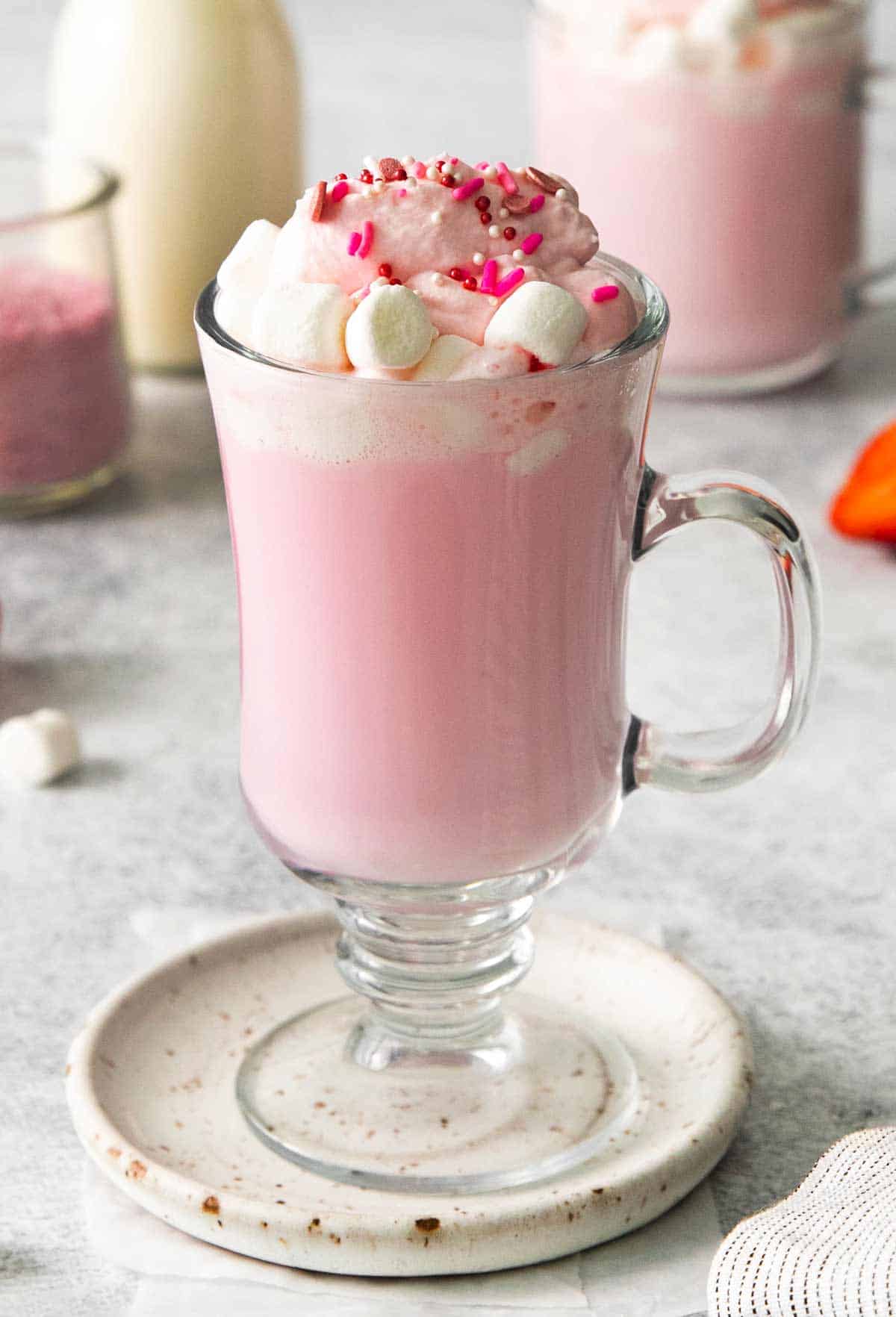 This is a photo of Hot Strawberry Milk served in a mug-style clear glass with a handle, topped with red and pink sprinkles and mini marshmallows. 